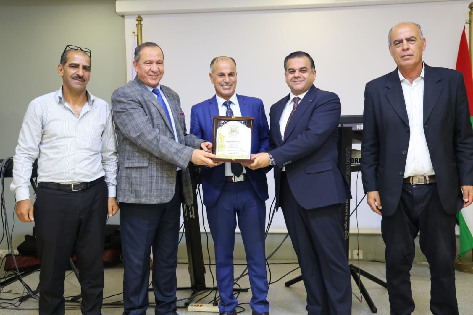 WhatsApp Image 2026-08-23 at 105938 PM - عروبة الاخباري | Oroba News Five men in suits stand together as one receives a ceremonial plaque on stage, with a flag in the background and a keyboard setup nearby in a formal event setting.