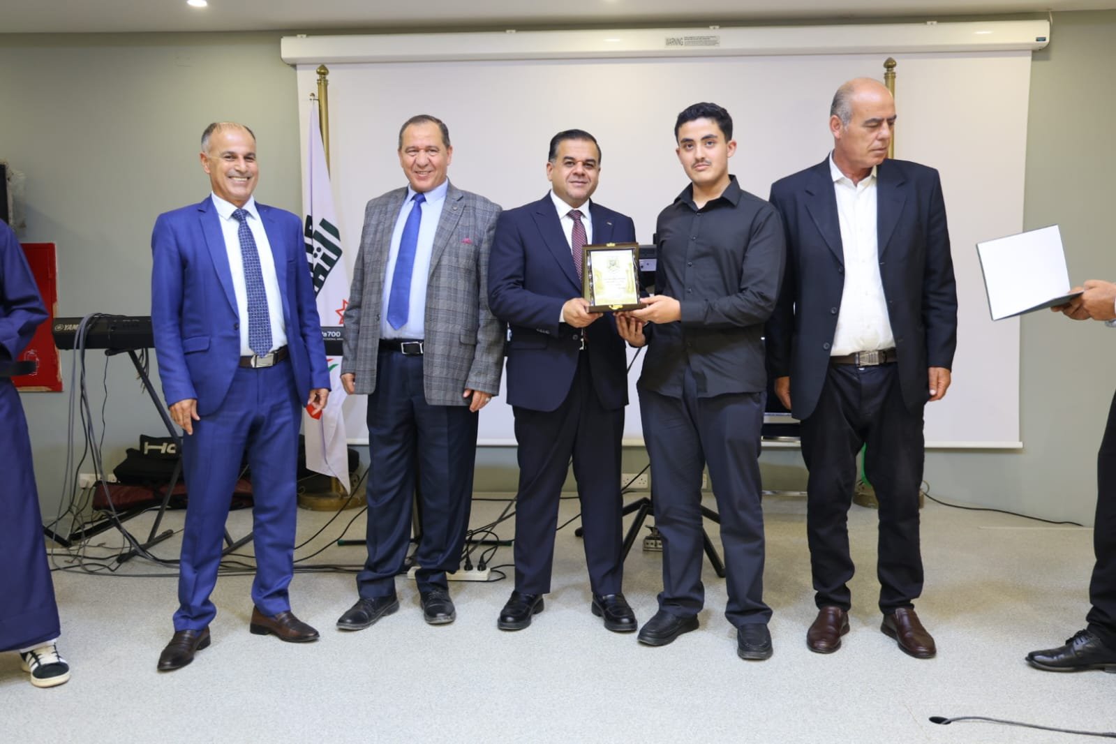 WhatsApp Image 2026-08-23 at 105955 PM - عروبة الاخباري | Oroba News Group of men in formal attire at an award ceremony, one handing a plaque to a younger man in black shirt and dark suit.