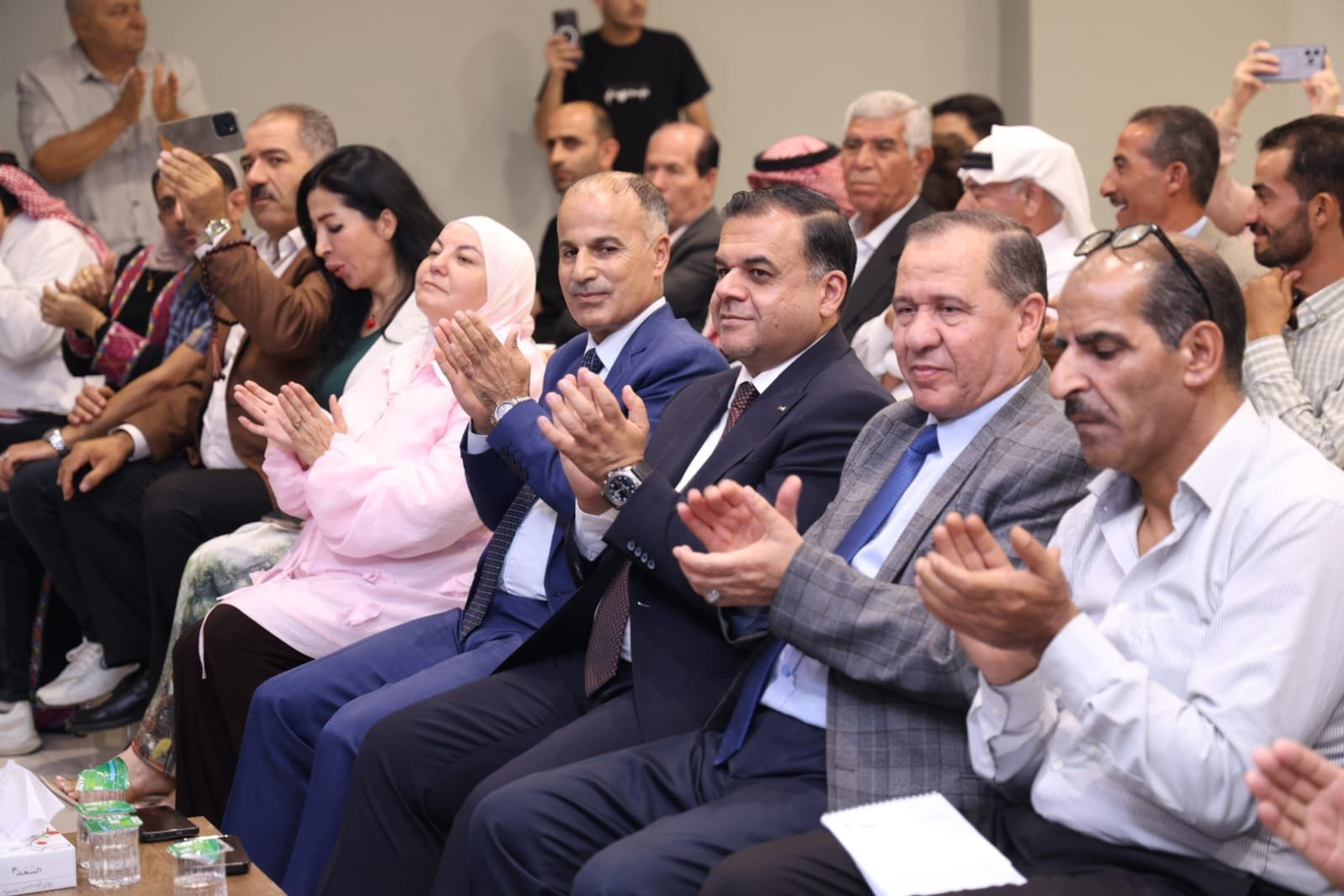WhatsApp Image 2026-08-23 at 105955 PM1 - عروبة الاخباري | Oroba News Group of formally dressed attendees sitting in rows, clapping during an indoor event; diverse mix of men and women including a woman in a pink hijab and others in suits and traditional attire.
