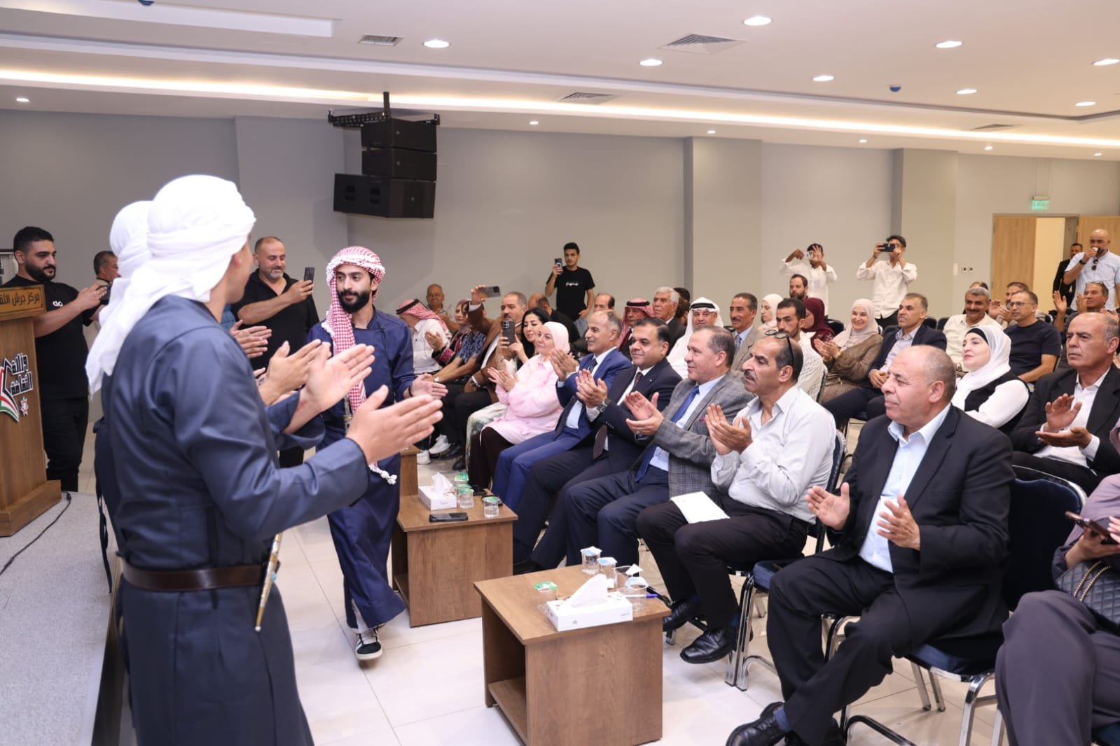 WhatsApp Image 2026-08-23 at 105955 PM2 - عروبة الاخباري | Oroba News Audience in a conference room applauds as two men in traditional dress stand at the front of a stage area.