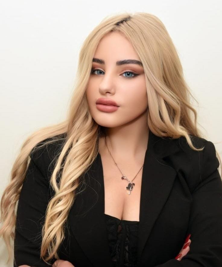 Portrait of a woman with long wavy blonde hair, wearing a black blazer and a pendant necklace, facing slightly to the left with neutral expression.