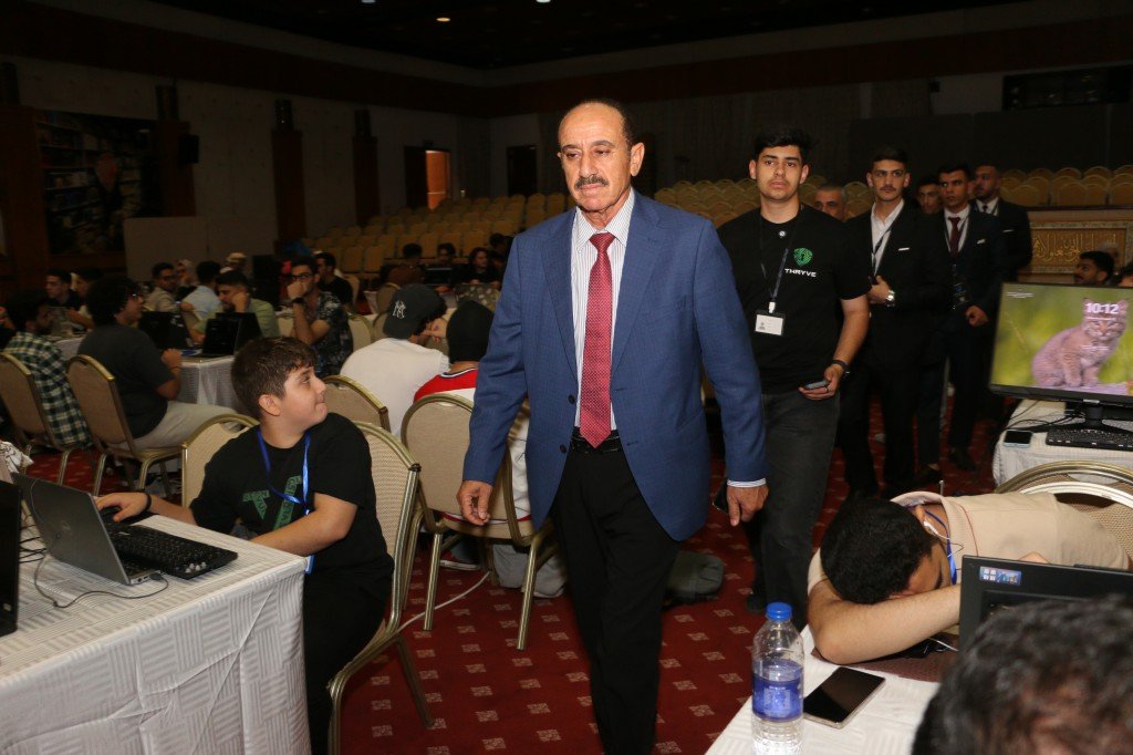 WhatsApp Image 2026-08-23 at 120306 AM2 - عروبة الاخباري | Oroba News Middle-aged man in a blue suit and red tie walks through a conference hall past attendees at tables with laptops nearby.