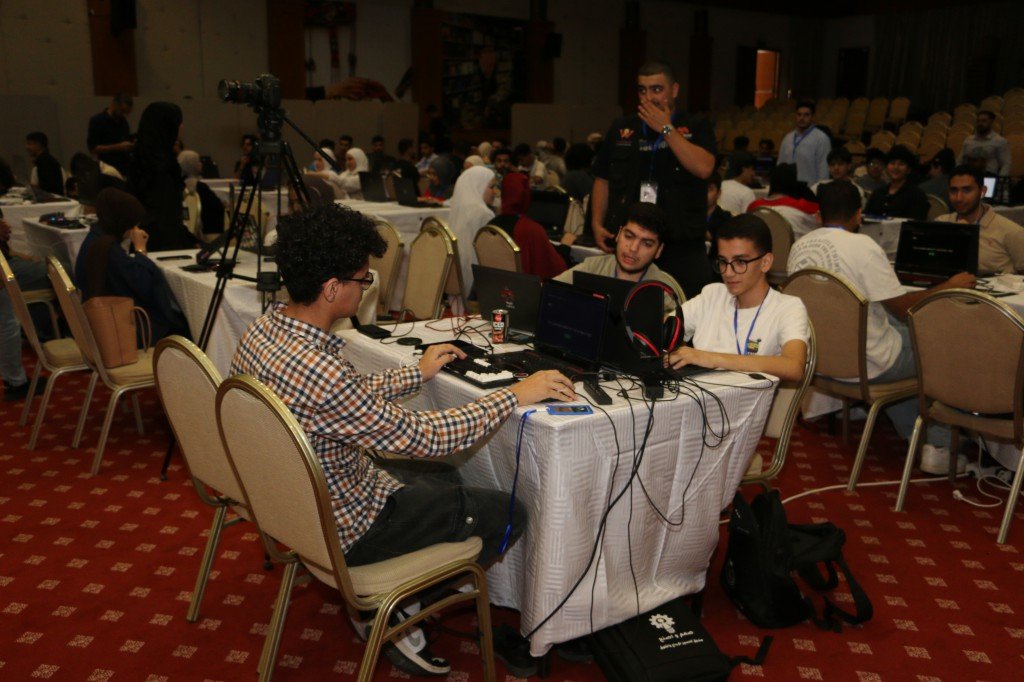 WhatsApp Image 2026-08-23 at 120307 AM - عروبة الاخباري | Oroba News Group of young people coding at a conference table with laptops; camera on tripod in background.