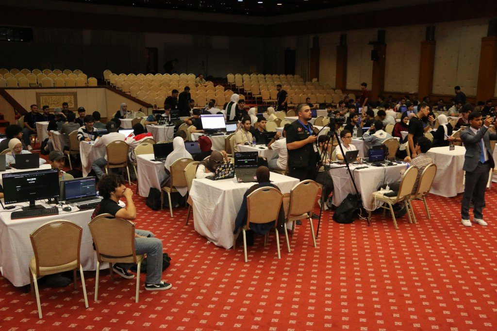 WhatsApp Image 2026-08-23 at 120307 AM1 - عروبة الاخباري | Oroba News Group of people coding on laptops at round tables in a large conference hall during a hackathon or tech event.