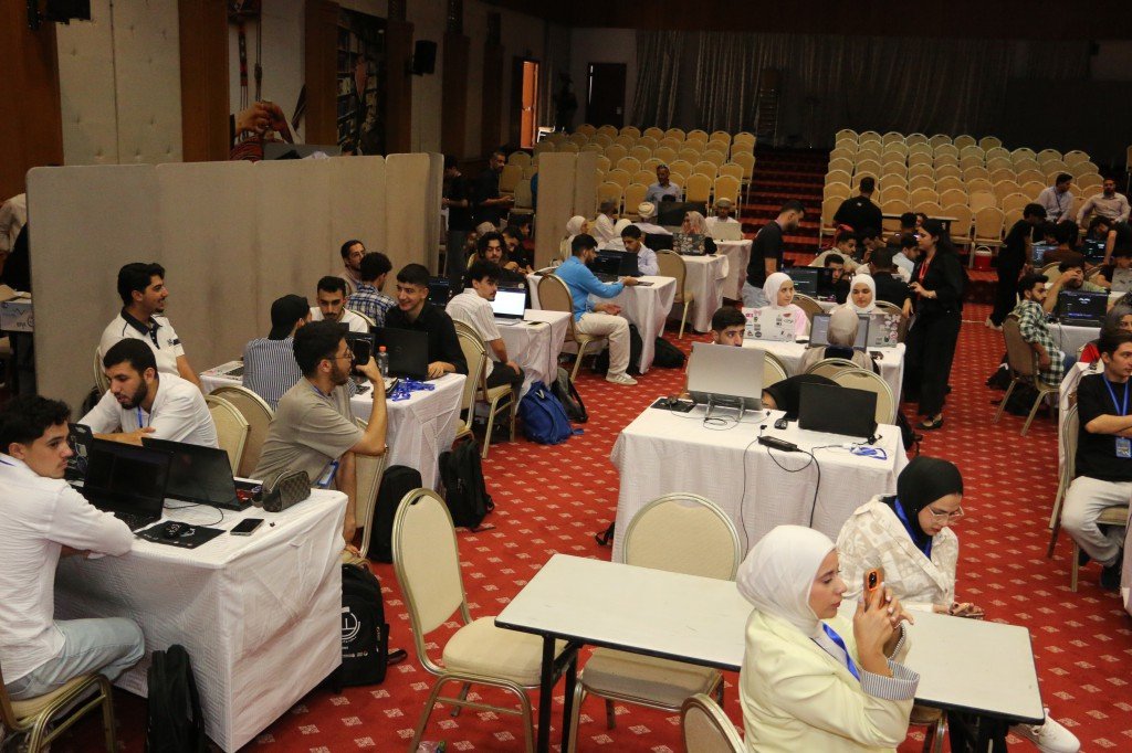 WhatsApp Image 2026-08-23 at 120307 AM2 - عروبة الاخباري | Oroba News Large hall with many people seated at tables coding on laptops during a hackathon or workshop.