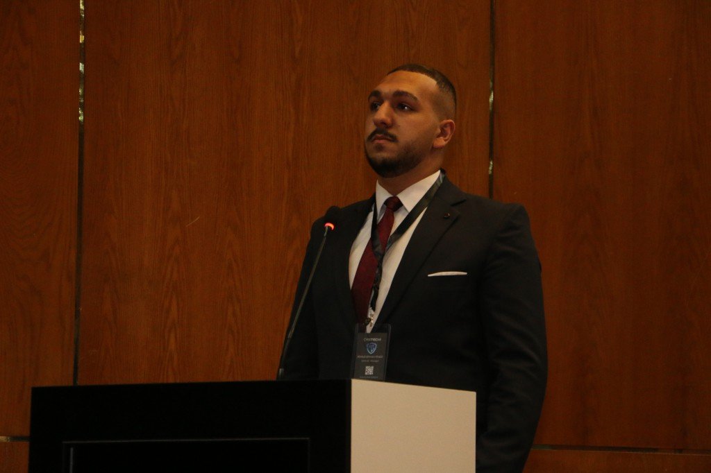 WhatsApp Image 2026-08-23 at 120308 AM - عروبة الاخباري | Oroba News Man in a black suit and maroon tie stands at a microphone behind a podium, speaking at a conference with wood-paneled walls behind him.