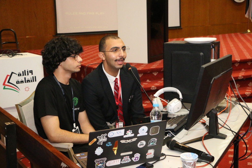 WhatsApp Image 2026-08-23 at 120312 AM - عروبة الاخباري | Oroba News Two men sit at a desk with laptops and microphones, presenting at a conference or event setup.
