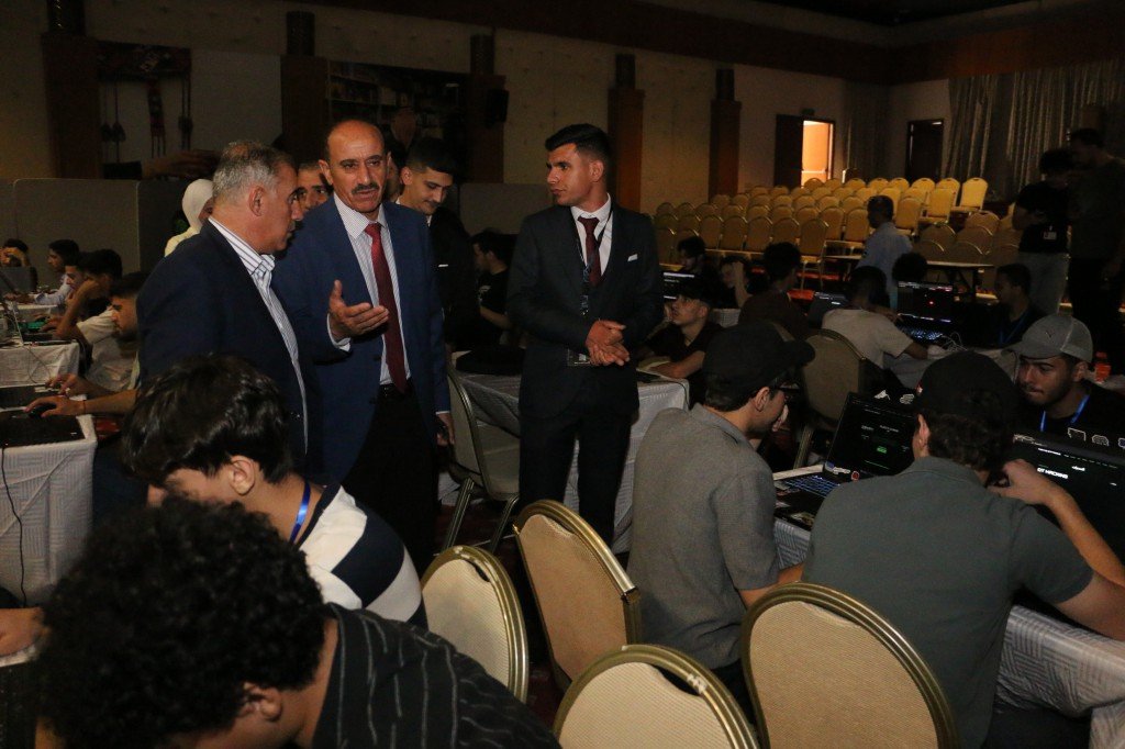 WhatsApp Image 2026-08-23 at 120314 AM - عروبة الاخباري | Oroba News Group of men in suits conversing with students at computers during a workshop in a large hall.