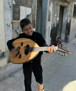 Boy in a black shirt plays a wooden oud on a city sidewalk, smiling as he strums the instrument.