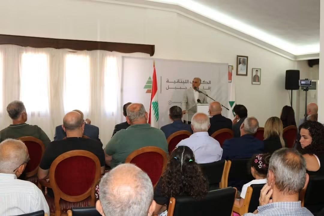 Speaker at a podium addressing a seated audience in a conference room; banner and Lebanese flags behind him.