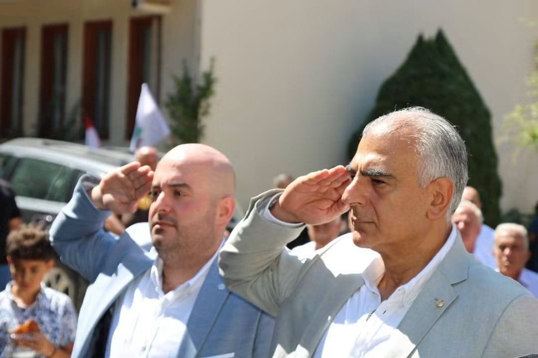 Two men in light suits salute during a formal outdoor ceremony, with onlookers behind them.