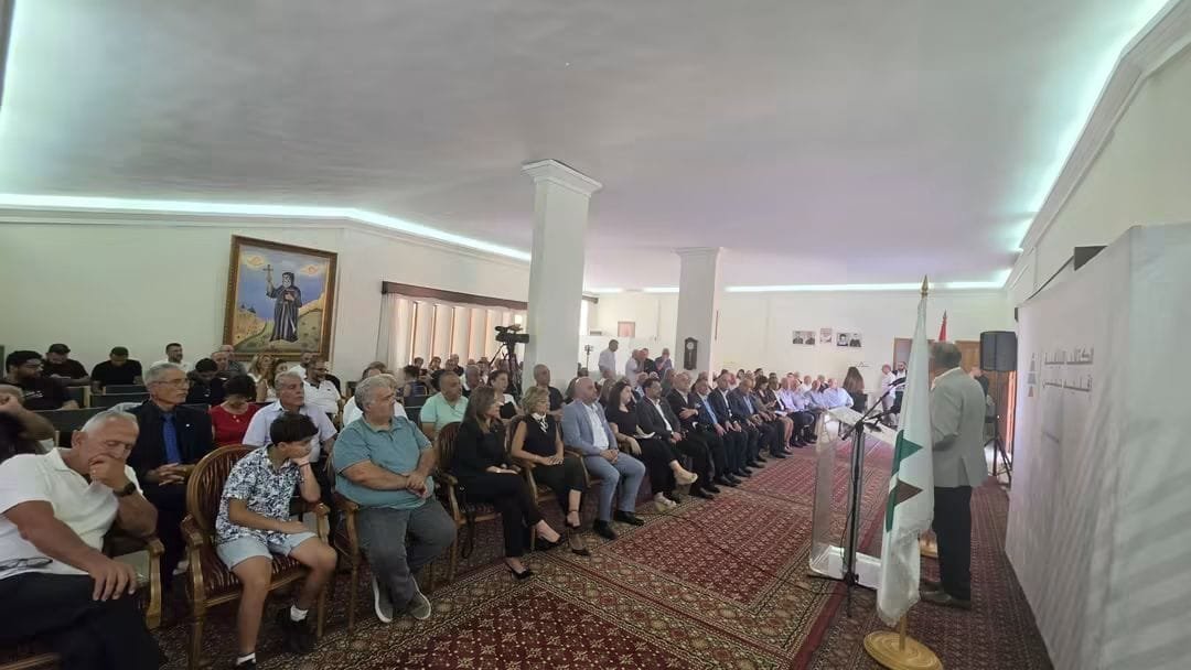 Audience seated in a conference room listening to a speaker at a podium with microphones, a painting on the wall behind them. 2) Attendees in formal attire facing a speaker in a carpeted hall. 3) Large crowd at a formal event or lecture, speaker at a lectern with a flag nearby. 4) Rows of people watching a presentation in a well-lit hall with a bannered wall to the right. 5) Indoor ceremony: people seated as a presenter speaks at a podium with a microphone.