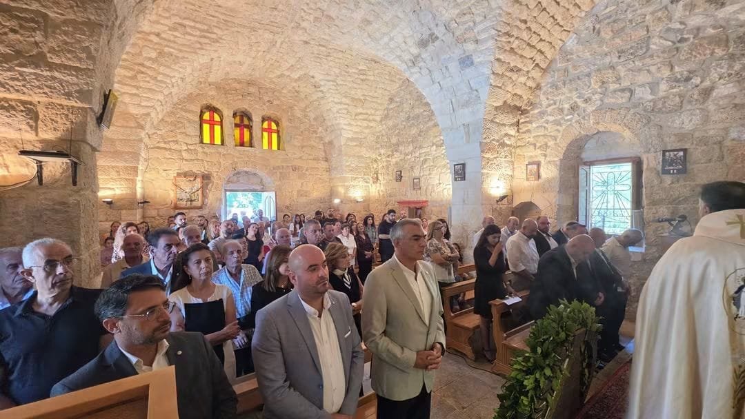 Crowded stone church interior during a ceremony; attendees sit in wooden pews as a priest in white robes leads near the front.