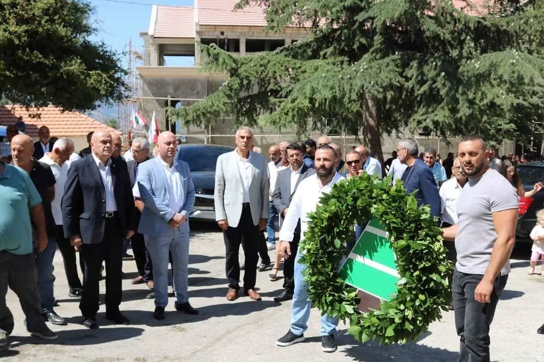Group of men in suits at an outdoor ceremony, with a large green wreath bearing a sign in the foreground.