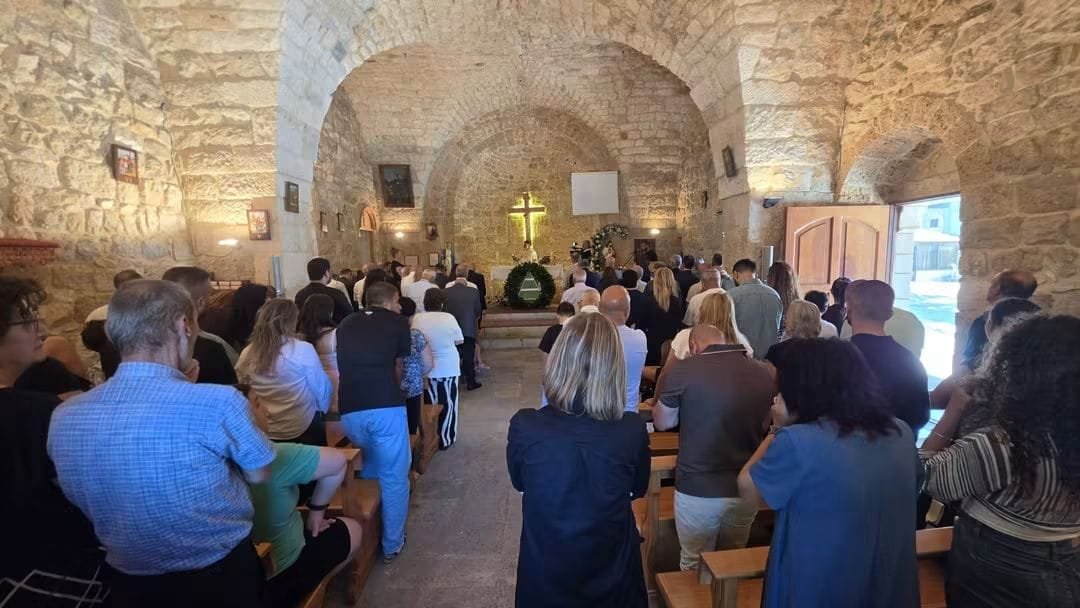 Inside a vaulted stone church, people sit on wooden pews facing the altar with a lit cross.