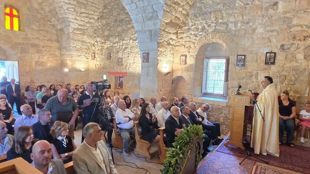 Priest in white robes delivers a sermon at a wooden pulpit in a vaulted stone church with seated and standing attendees.