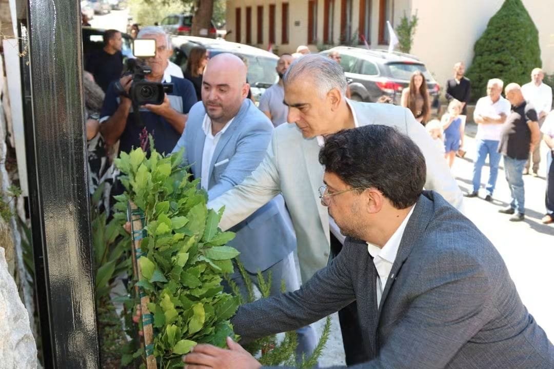 Men in suits lay a green wreath at a memorial as onlookers and photographers watch.
