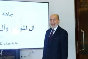 Man in a dark suit and tie stands beside a large screen displaying Arabic text in a conference room setting.