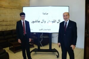 Two men in suits pose beside a large screen displaying Arabic text in a conference room setting.