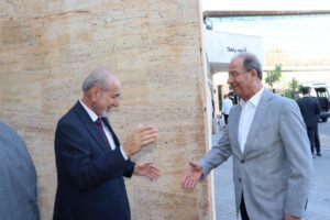 Two men in business attire shake hands and smile outdoors beside a beige stone wall.