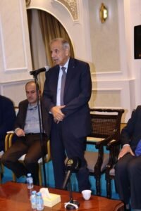 A suited older man stands at a microphone delivering a speech in a formal meeting room, with seated colleagues nearby.