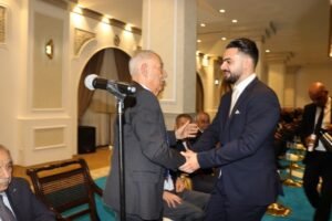 Older man in a gray suit shakes hands with a younger man in a navy suit during a formal ceremony, microphone nearby, audience seated behind.