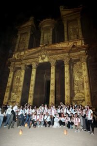 Group of people in white shirts posing in front of a tall, ornate ancient facade lit at night with lanterns on the ground.