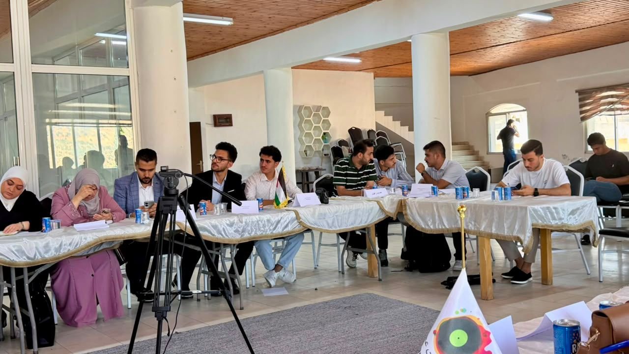 Panel of people seated along a long, covered conference table in a bright room, with a video camera on a tripod in the foreground.