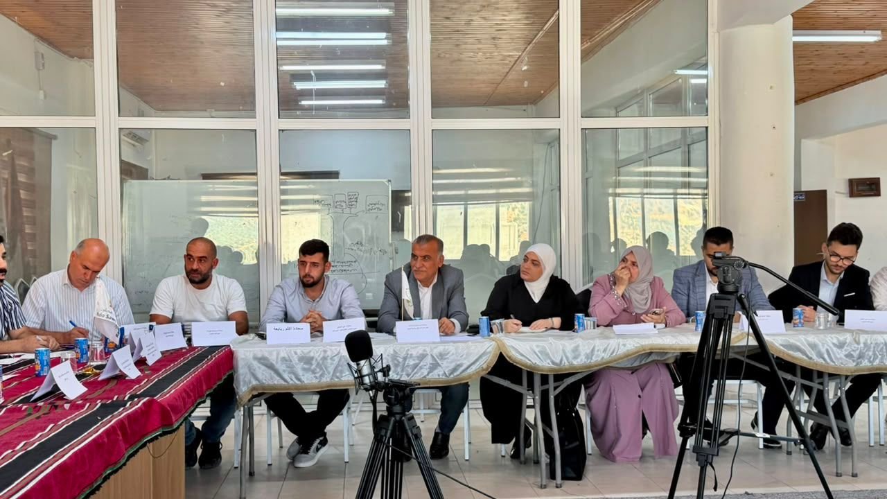 Panel discussion with nine people seated along a long table in a bright room, microphones and nameplates in front of them.