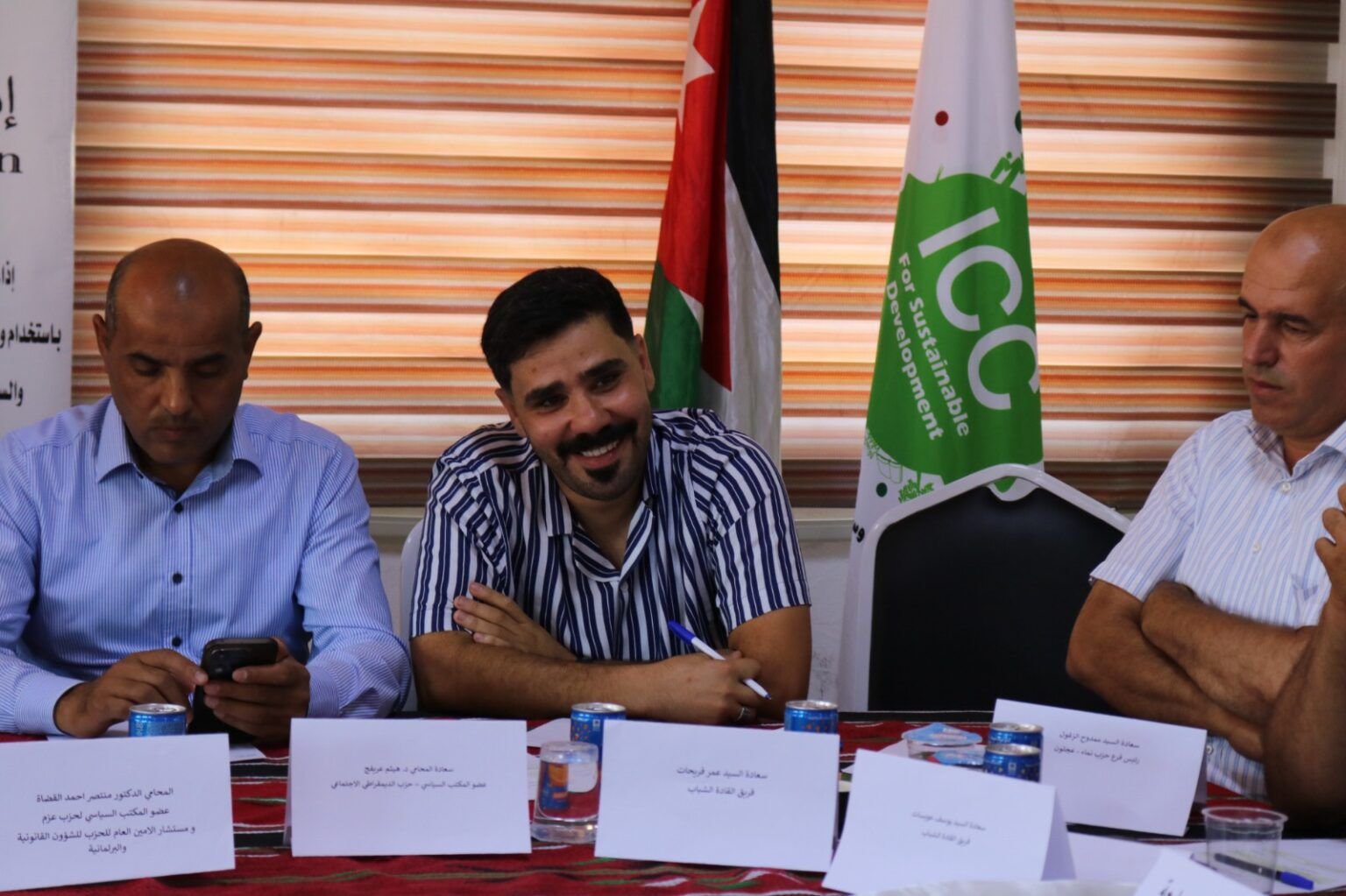 Three men sit at a conference table with name cards, the center man smiling, left man looking at his phone; striped blinds and flags behind them.