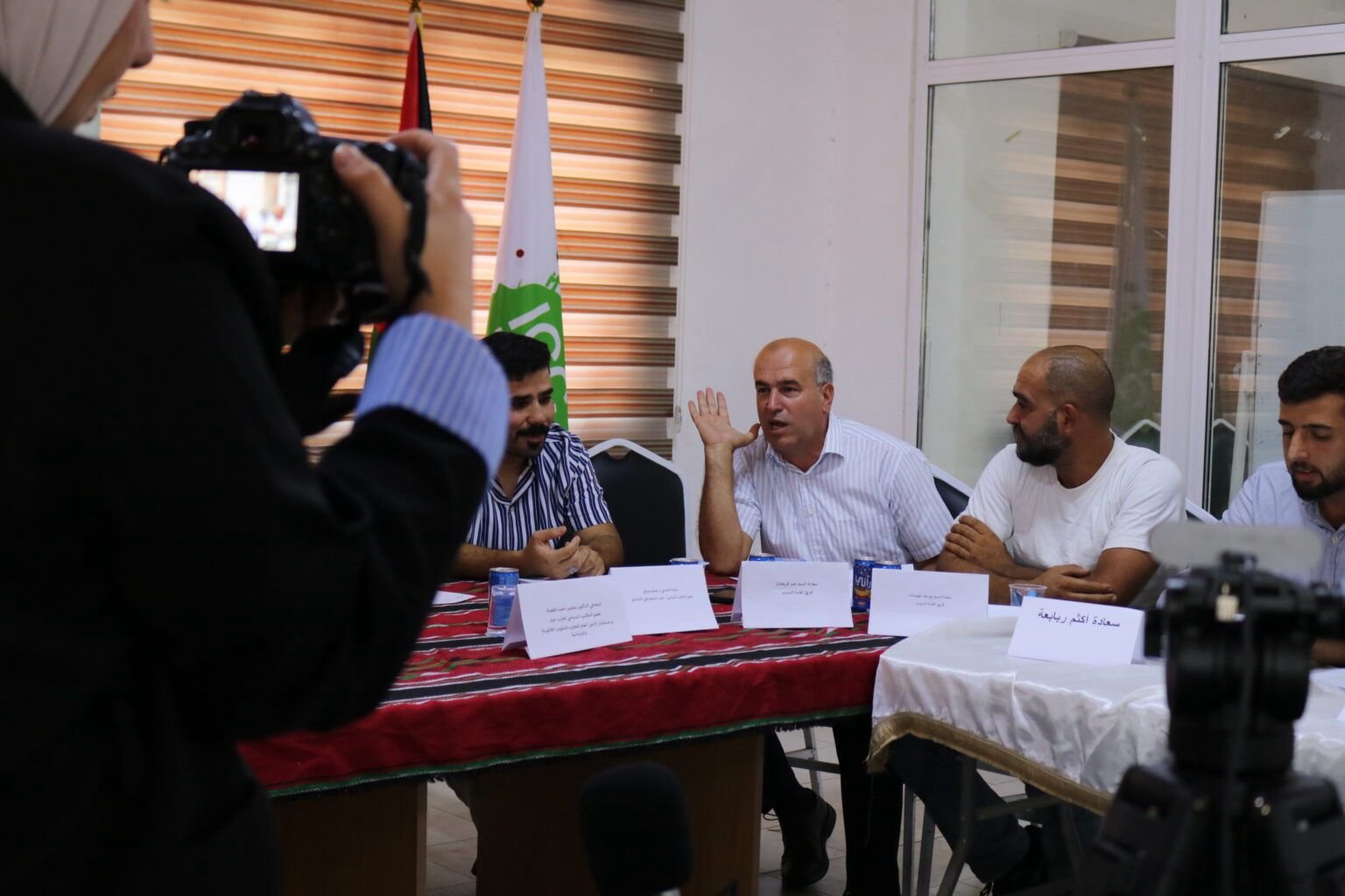 Panel of men seated at a long table with nameplates, a man raises his hand while speaking as a camera operator films from the left.
