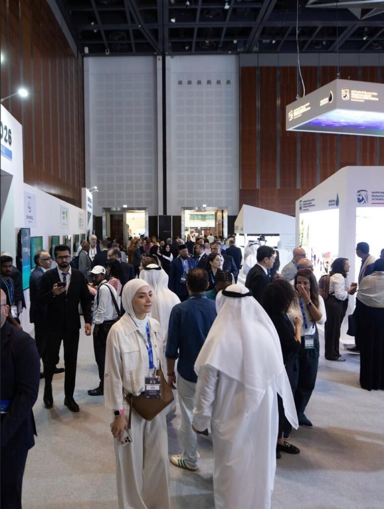 Crowded exhibition hall with attendees wearing badges, some in white traditional robes and business attire, browsing booths along white partition walls.