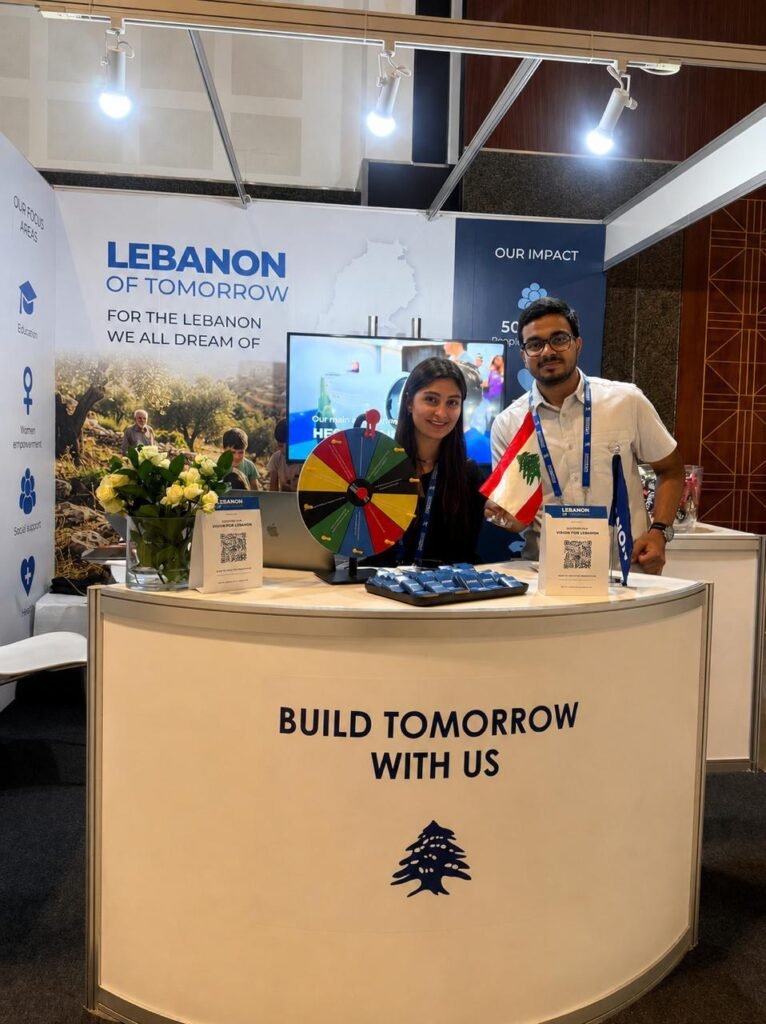 Two exhibitors stand behind a curved booth at a Lebanon-themed expo, with a colorful prize wheel and Lebanon flags on display.