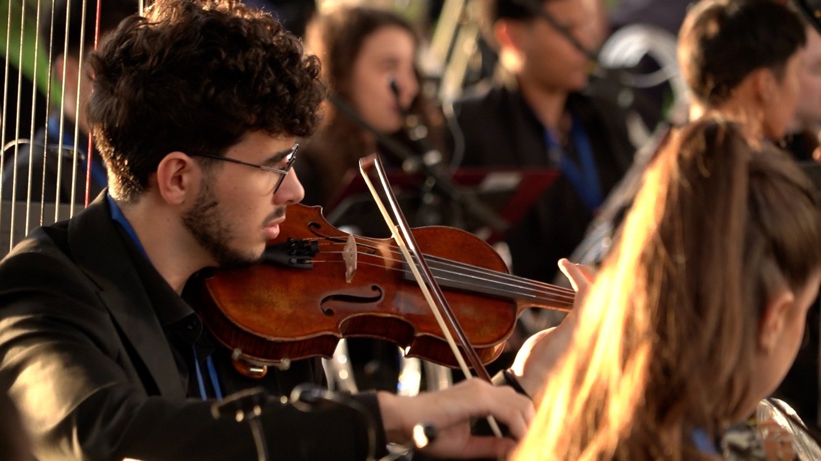 WhatsApp Image 2026-08-29 at 123200 PM 2 - عروبة الاخباري | Oroba News Young man with dark hair and glasses plays a violin in a small ensemble, with background musicians blurred.
