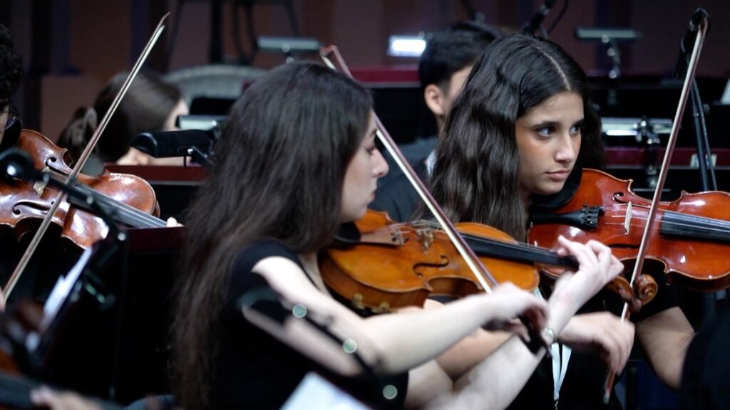 WhatsApp Image 2026-08-29 at 123200 PM 3 - عروبة الاخباري | Oroba News Two young women play violins in an orchestra, concentrating on their performance amid other musicians in the background.