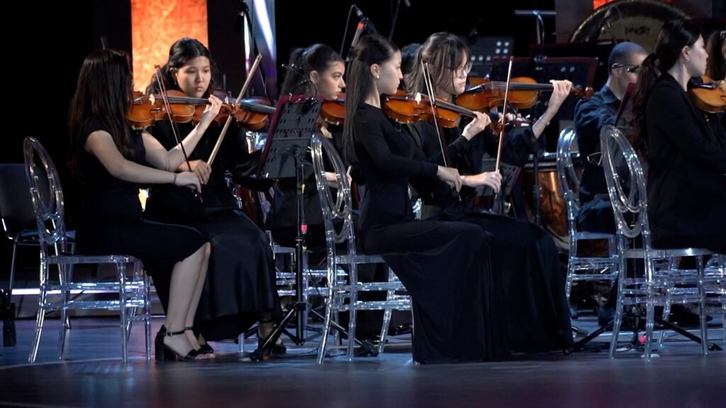 WhatsApp Image 2026-08-29 at 123200 PM 4 - عروبة الاخباري | Oroba News Female violinists in black dresses seated on transparent chairs, performing on a concert stage.