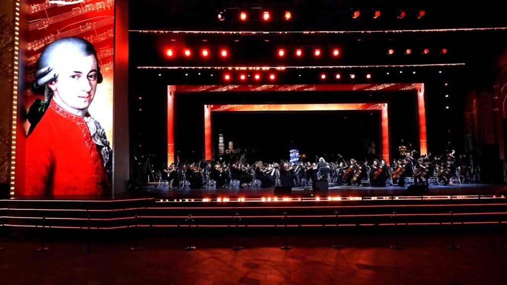 WhatsApp Image 2026-08-29 at 123200 PM 5 - عروبة الاخباري | Oroba News Orchestra performing on a red-lit stage with a towering portrait of a man in a red coat on the left.