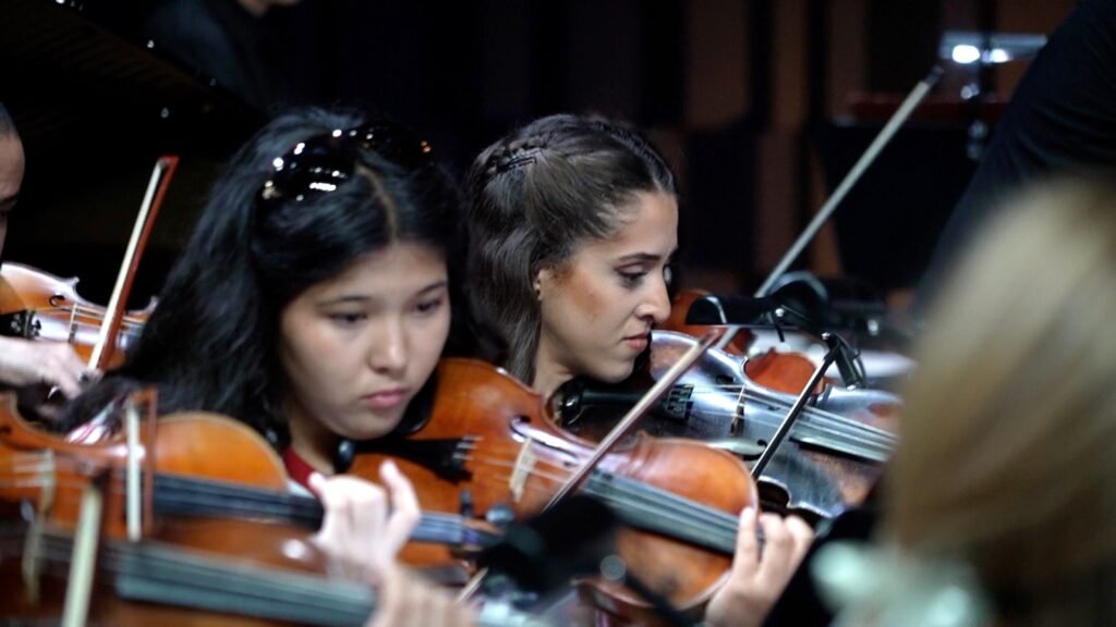 WhatsApp Image 2026-08-29 at 123200 PM 6 - عروبة الاخباري | Oroba News Two young women violinists focused on their instruments in an orchestra performance.