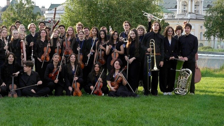 WhatsApp Image 2026-08-29 at 123201 PM 1 - عروبة الاخباري | Oroba News Large group of young musicians in black outfits posing with violins, cellos, and brass instruments on a grassy outdoor lawn.