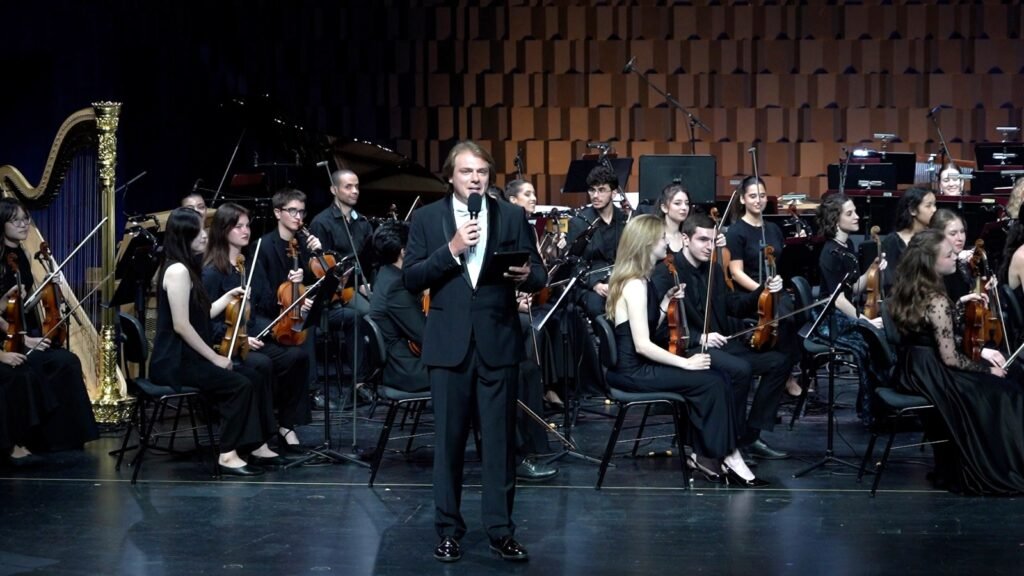 WhatsApp Image 2026-08-29 at 123201 PM - عروبة الاخباري | Oroba News Conductor in a tuxedo holding a microphone, addressing the audience on stage with a full orchestra behind him