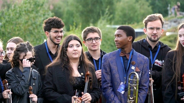 WhatsApp Image 2026-08-29 at 123201 PM 3 - عروبة الاخباري | Oroba News Group of young musicians outdoors, some with violins and a trombone, wearing lanyards at a daytime event.