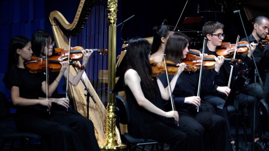 WhatsApp Image 2026-08-29 at 123201 PM 4 - عروبة الاخباري | Oroba News Group of young musicians in black playing violins on stage, with a harp visible in the background.