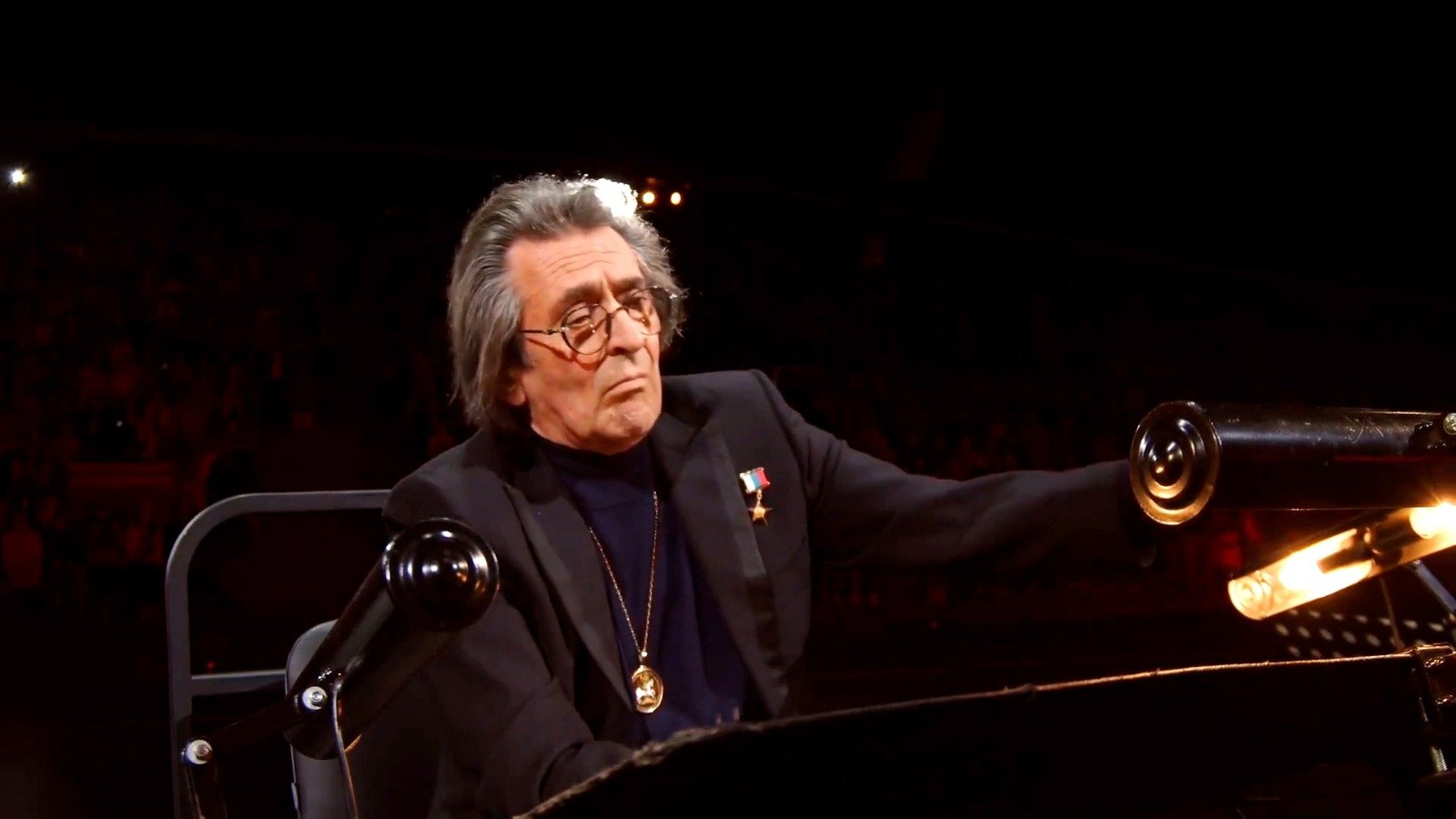 WhatsApp Image 2026-08-29 at 123202 PM 4 - عروبة الاخباري | Oroba News Older man with gray hair and glasses sits at a piano on a dimly lit stage, wearing a dark suit and medal while playing or preparing to play.