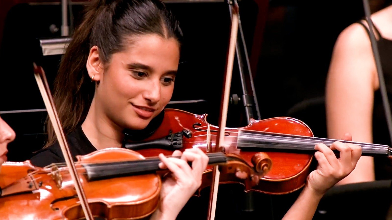 WhatsApp Image 2026-08-29 at 123202 PM 5 - عروبة الاخباري | Oroba News Young woman smiling while playing a violin in a group performance on stage.