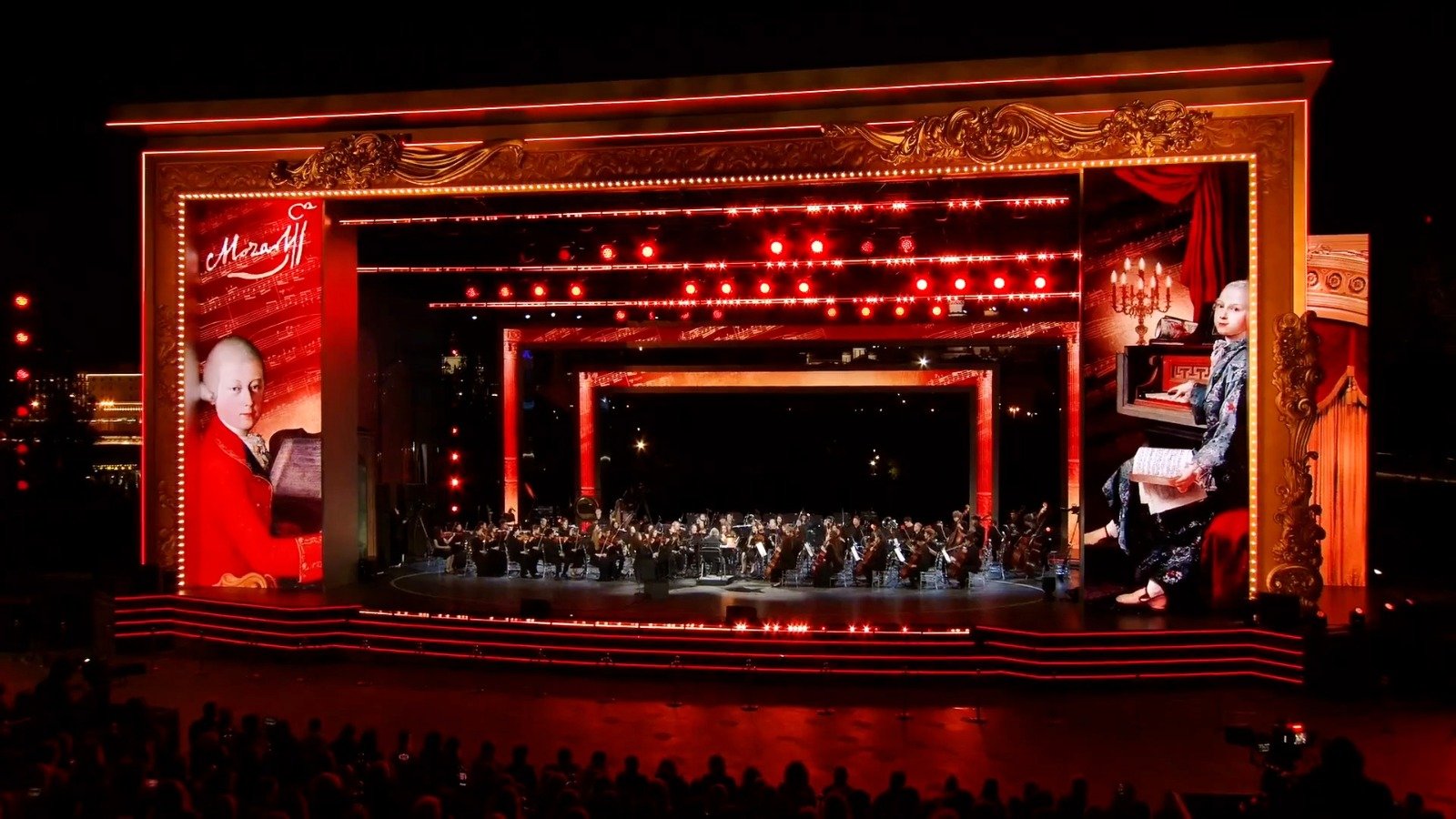 WhatsApp Image 2026-08-29 at 123202 PM 6 - عروبة الاخباري | Oroba News Orchestra performing on a lit, ornate red-framed stage at night, audience silhouettes in foreground.