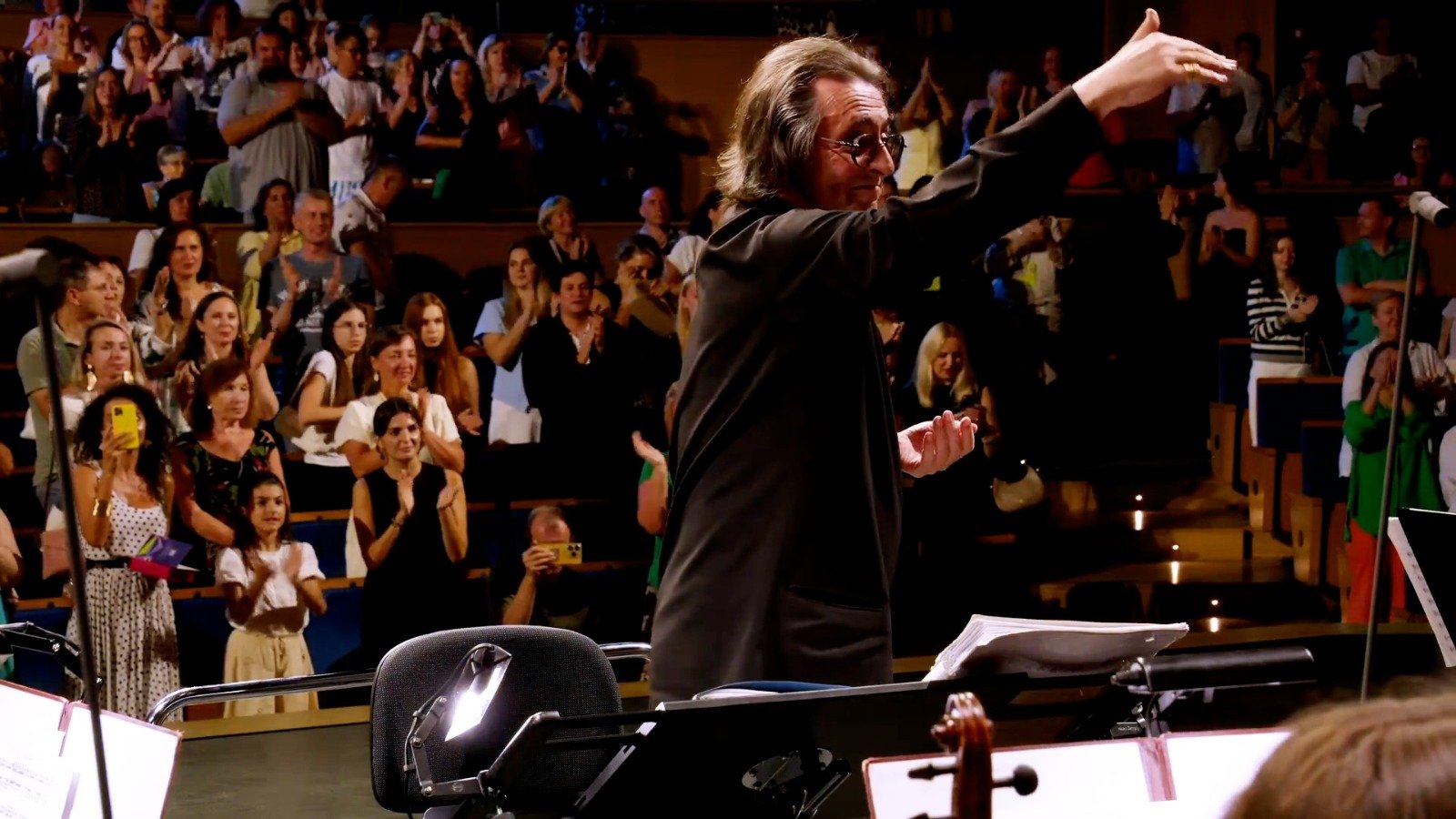 WhatsApp Image 2026-08-29 at 123203 PM 2 - عروبة الاخباري | Oroba News Conductor on stage raising a baton to lead a performance as the audience applauds in the theater hall.