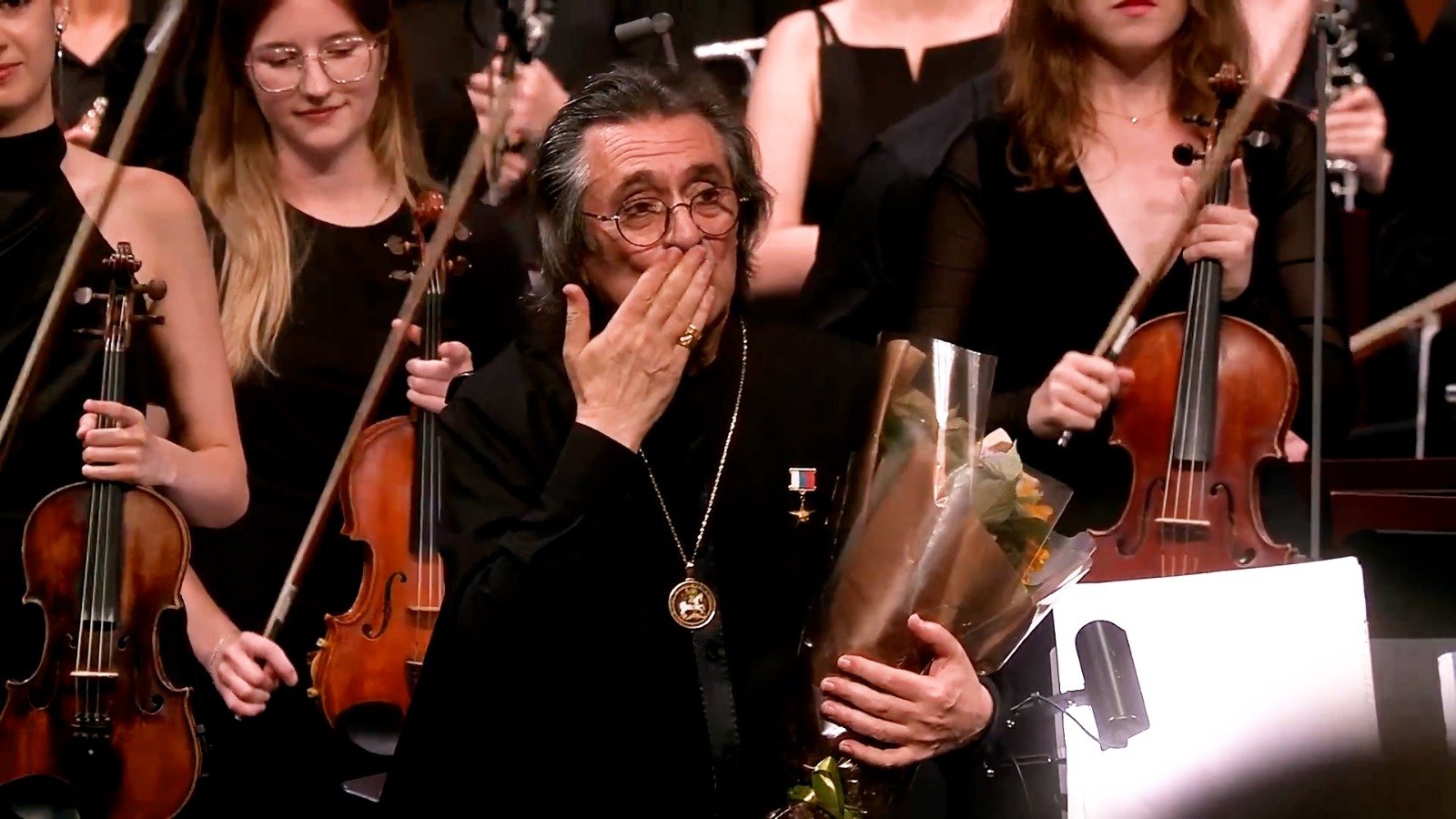 WhatsApp Image 2026-08-29 at 123203 PM - عروبة الاخباري | Oroba News Elderly musician in black with a medal, holding flowers and covering his mouth in surprise as violinists stand behind him.