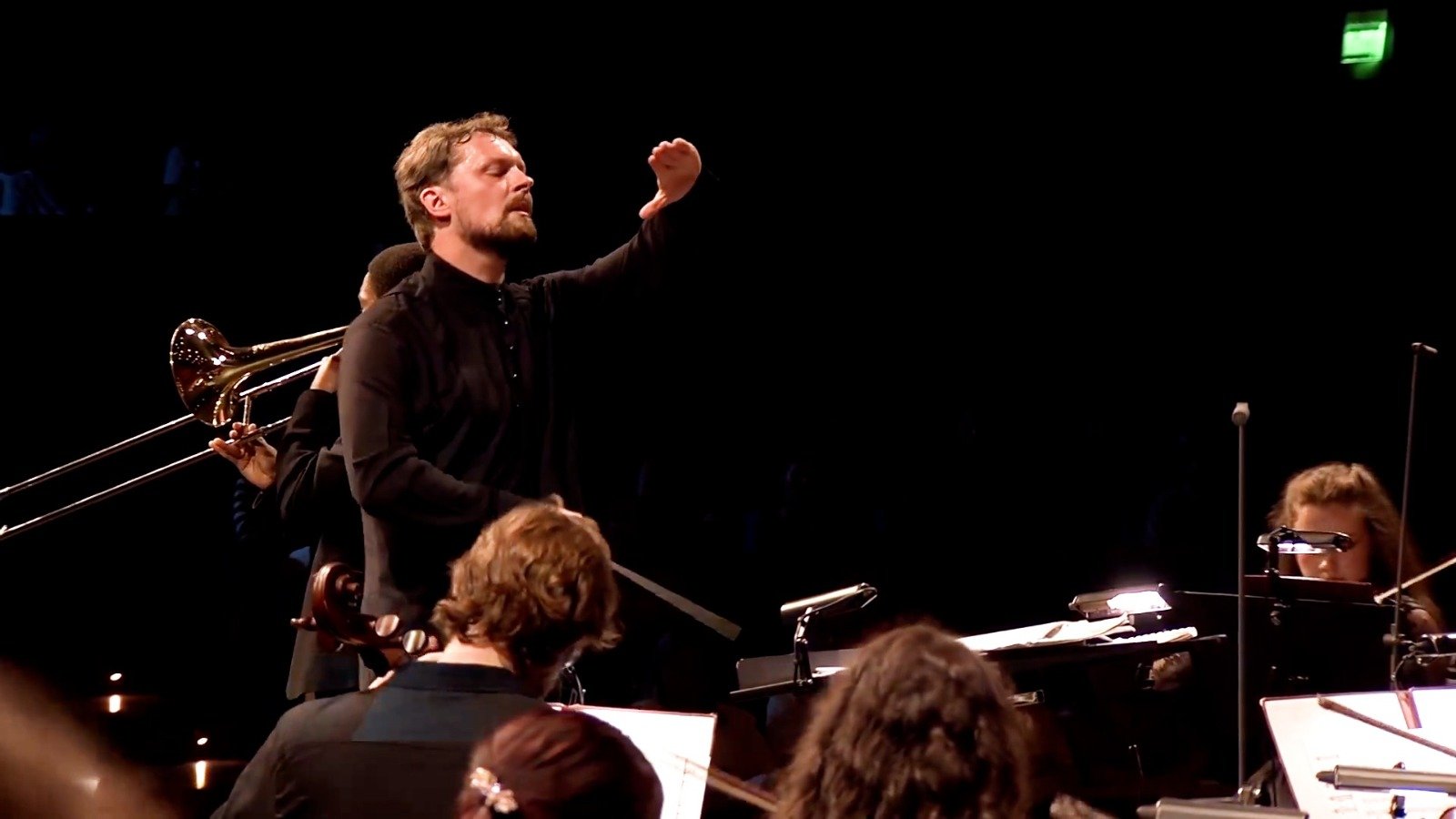 WhatsApp Image 2026-08-29 at 123204 PM 1 - عروبة الاخباري | Oroba News Conductor raises his right hand while leading a small orchestra on stage; trombone visible on the left and musicians read sheet music.
