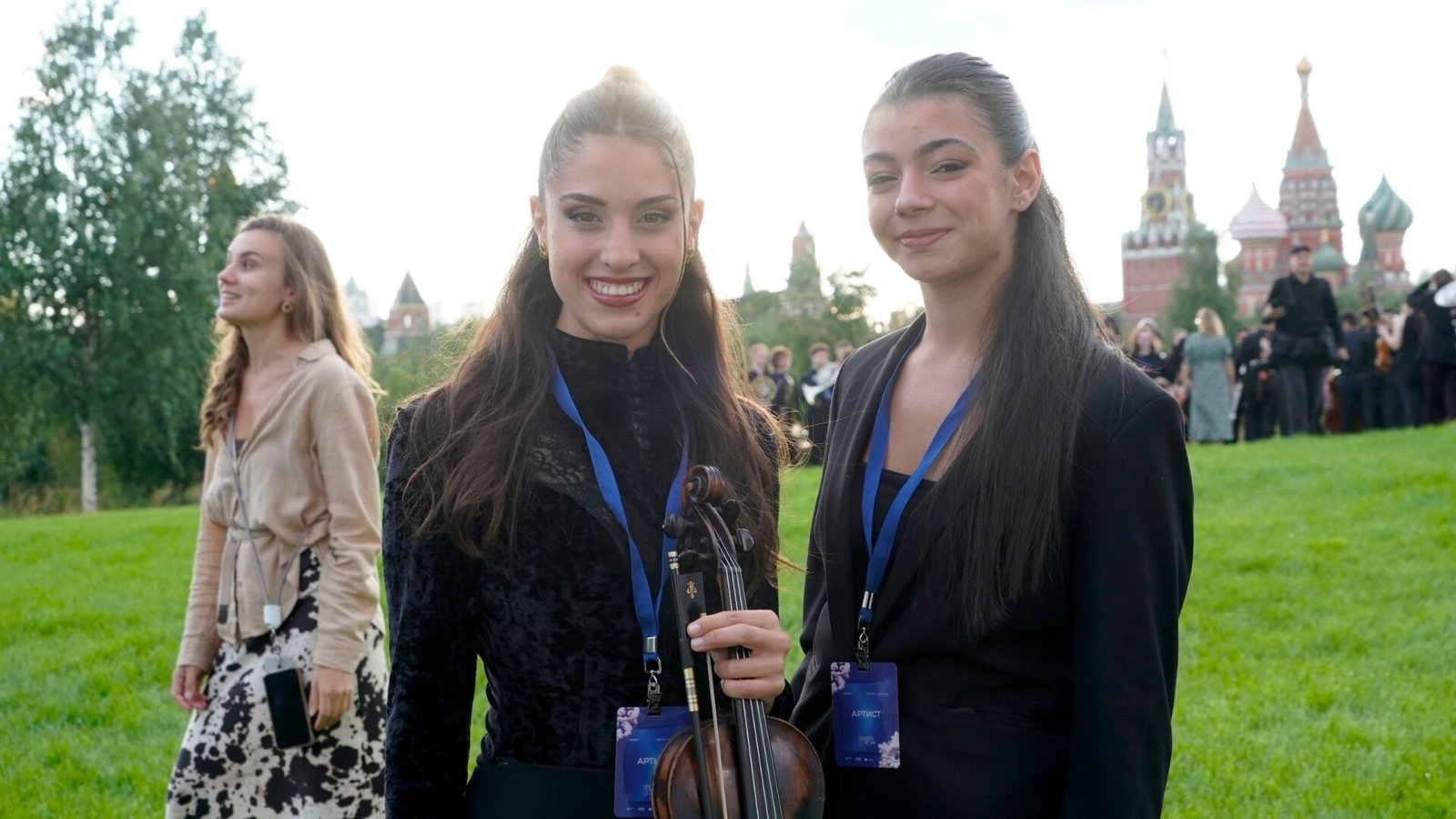 WhatsApp Image 2026-08-29 at 123204 PM 2 - عروبة الاخباري | Oroba News Two smiling women in formal attire with a violin and bow, wearing blue lanyards, outdoors on a green lawn with distant towers in the background.