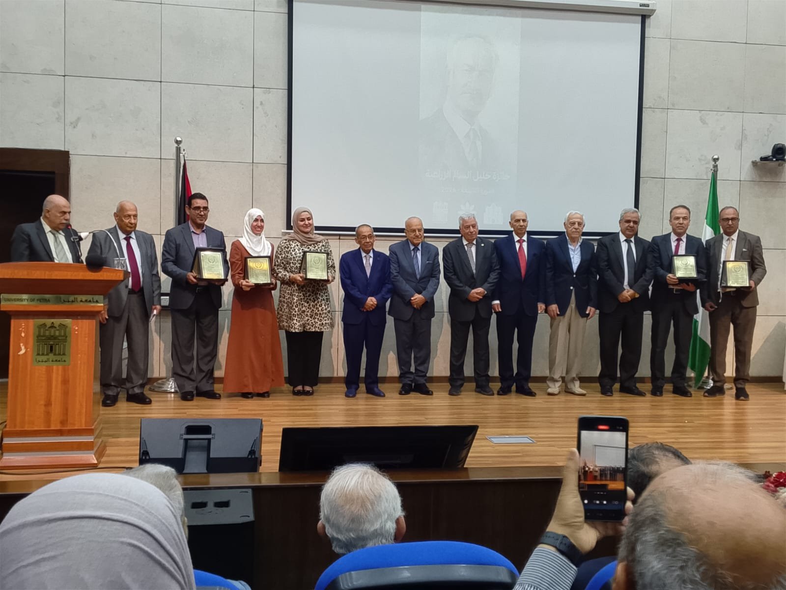 WhatsApp Image 2026-08-29 at 43332 PM - عروبة الاخباري | Oroba News Group of formally dressed men and women on a stage holding framed awards during a ceremony at a university.